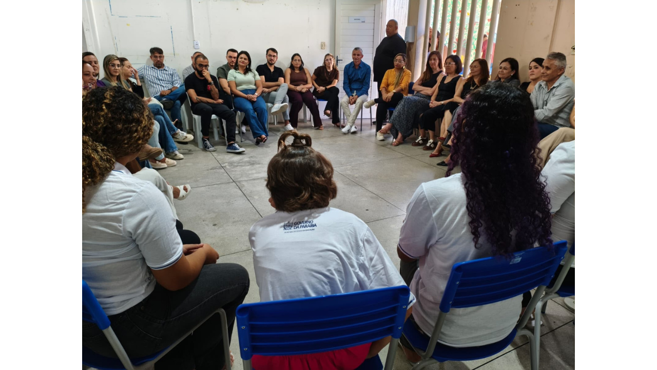 Cerca de vinte pessoas, com vestimentas casuais, estão sentadas em cadeiras dispostas em um grande círculo. Em primeiro plano, estão três participantes de costas, vestindo camisetas brancas. A pessoa do meio tem o cabelo nos ombros, com uma parte do cabelo presa, e veste uma camisa com o logotipo "Governo da Paraíba - Secretaria do Estado da Educação". As paredes da sala são brancas e o piso cinza. Ao fundo, há uma porta e uma janela com grades.