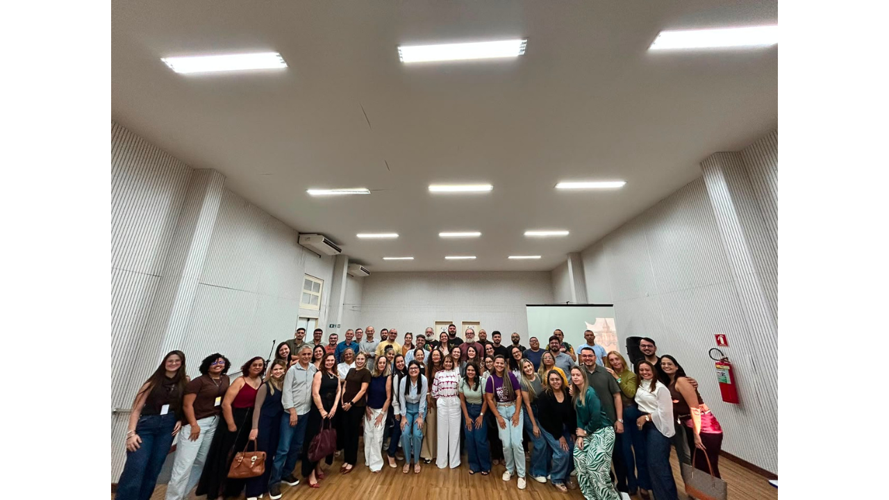Em um auditório bem iluminado, um grande grupo multirracial de aproximadamente quarenta pessoas posa de pé para a câmera. O grupo veste trajes variados, com predominância de calças jeans, camisetas coloridas e camisas. Ao fundo, as paredes são brancas e têm isolamento acústico ripado. A sala tem um teto alto com lâmpadas fluorescentes embutidas e uma tela de projeção branca à direita, apagada. O piso é de madeira clara.