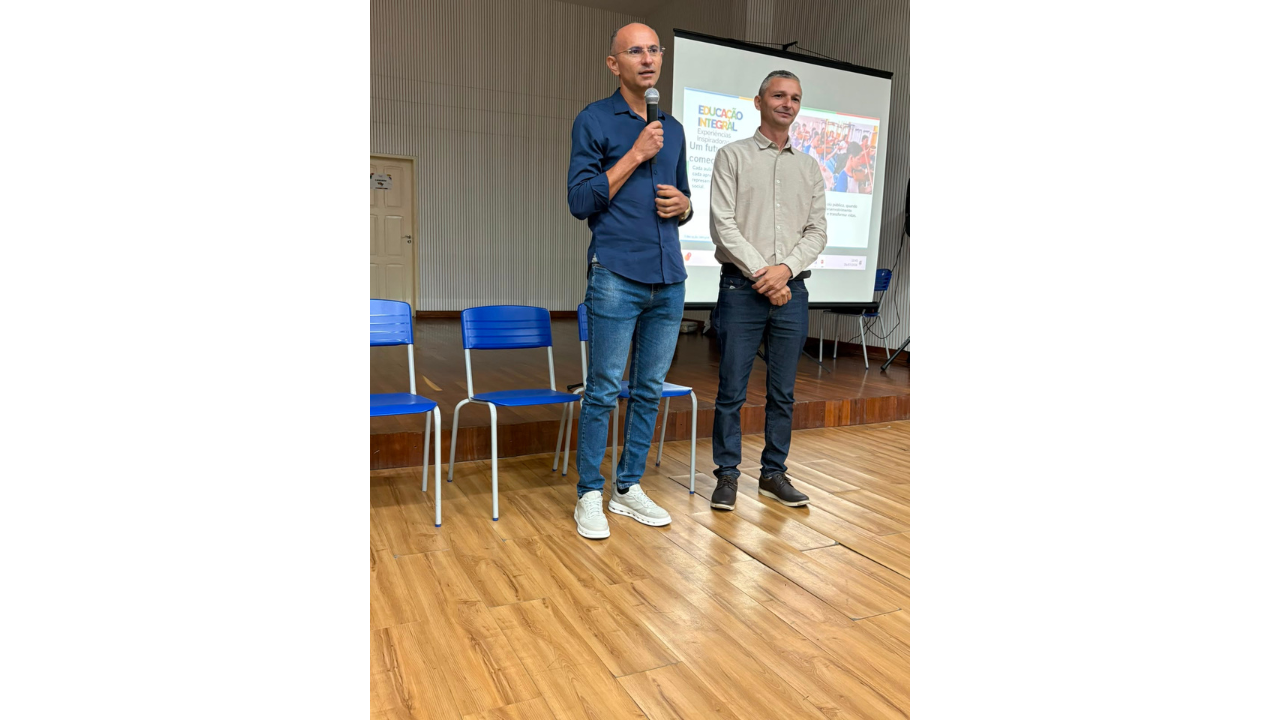 Dois homens fazem uma apresentação em uma sala com piso de madeira. O homem da esquerda, vestindo camisa azul escura e óculos, segura o microfone e fala. O homem da direita, de camisa bege, ouve com as mãos cruzadas à frente. Ao fundo, uma tela de projeção com o texto "Educação Integral" e três cadeiras plásticas azuis vazias.