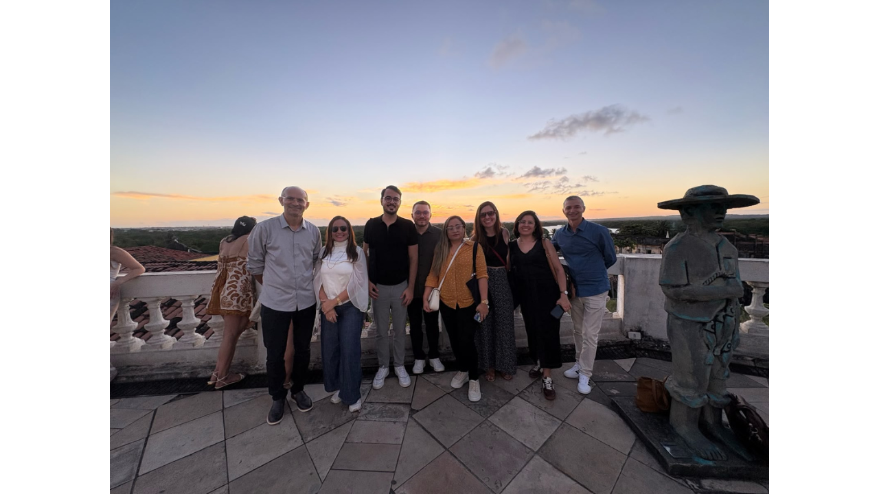 Um grupo de oito adultos posa em pé em um terraço de pedra ao pôr do sol. O céu tem cores laranja e azul. O grupo sorri para a câmera com vestimentas casuais. À direita, uma escultura de pedra escura de uma figura humana com chapéu. Atrás das pessoas, há um guarda-corpo em pedra na cor branca.
