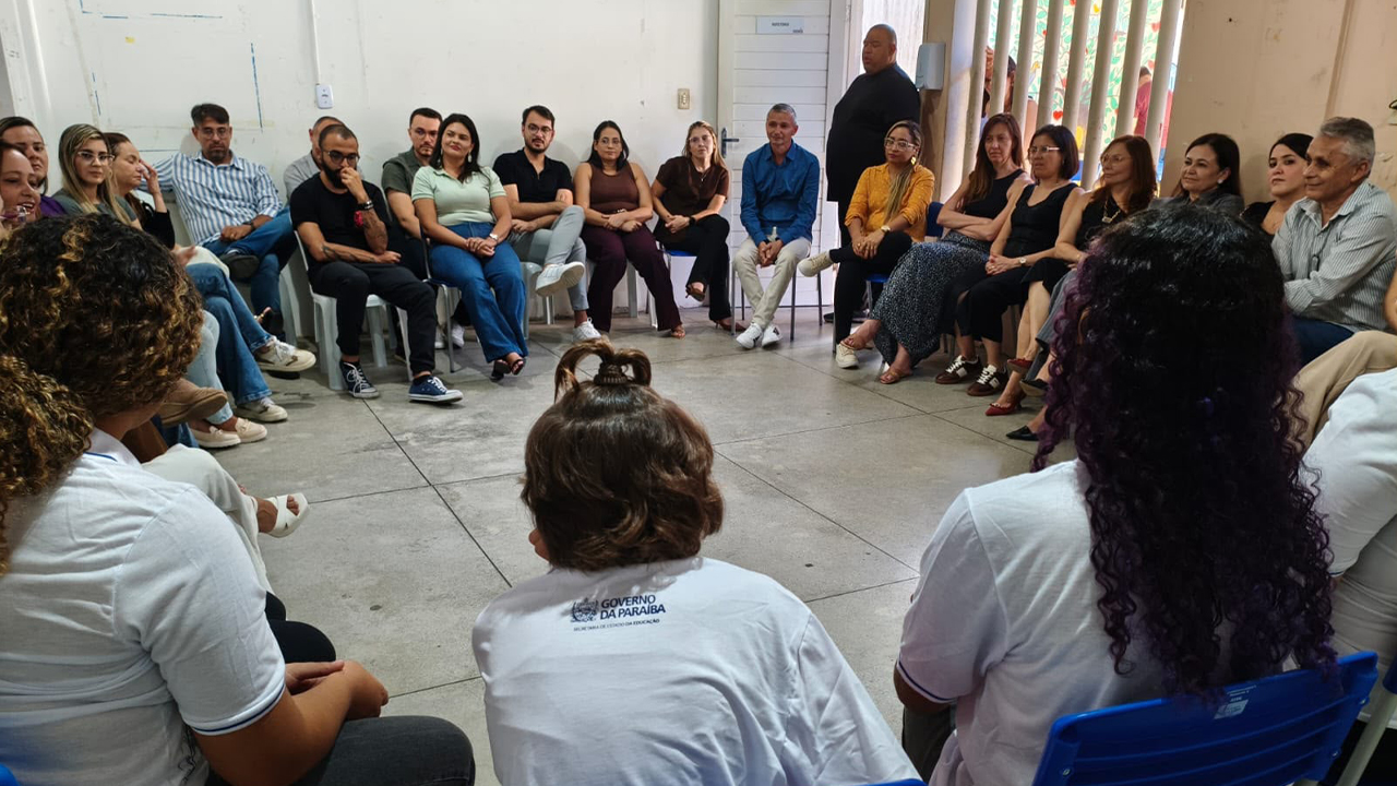 Cerca de vinte pessoas, com vestimentas casuais, estão sentadas em cadeiras dispostas em um grande círculo. Em primeiro plano, estão três participantes de costas, vestindo camisetas brancas. A pessoa do meio tem o cabelo nos ombros, com uma parte do cabelo presa, e veste uma camisa com o logotipo "Governo da Paraíba - Secretaria do Estado da Educação". As paredes da sala são brancas e o piso cinza. Ao fundo, há uma porta e uma janela com grades.