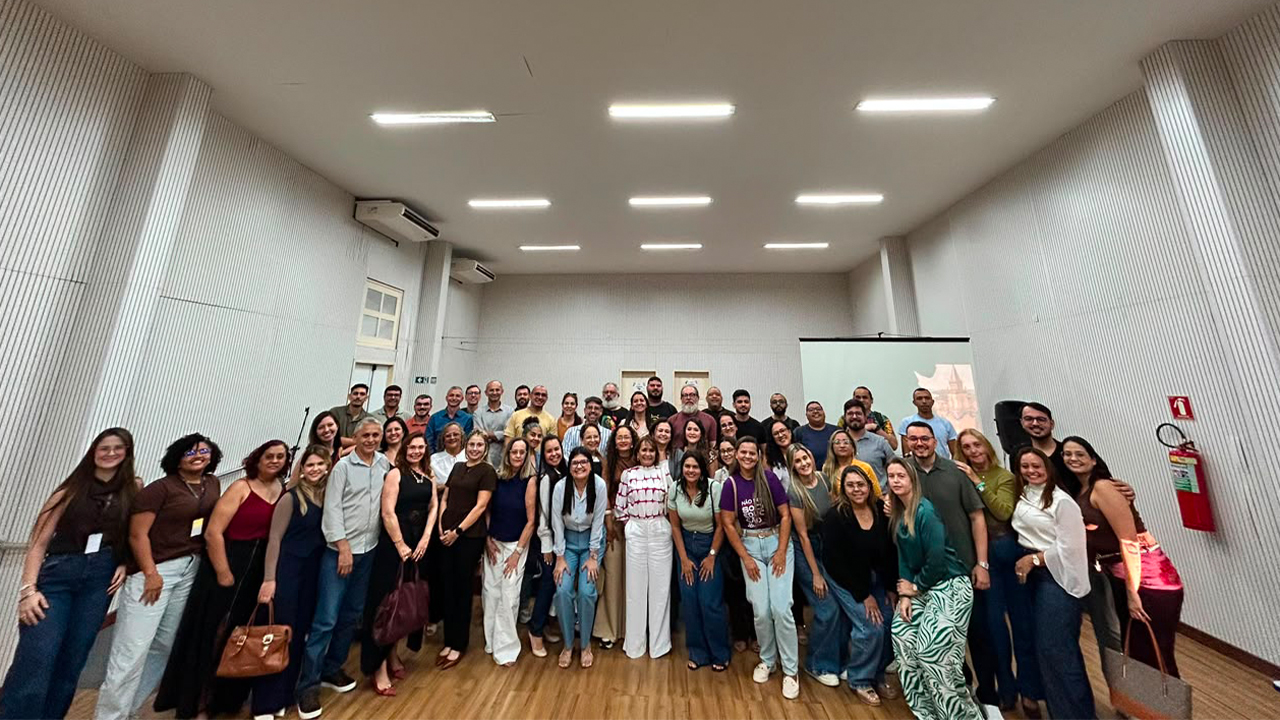 Em um auditório bem iluminado, um grande grupo multirracial de aproximadamente quarenta pessoas posa de pé para a câmera. O grupo veste trajes variados, com predominância de calças jeans, camisetas coloridas e camisas. Ao fundo, as paredes são brancas e têm isolamento acústico ripado. A sala tem um teto alto com lâmpadas fluorescentes embutidas e uma tela de projeção branca à direita, apagada. O piso é de madeira clara.