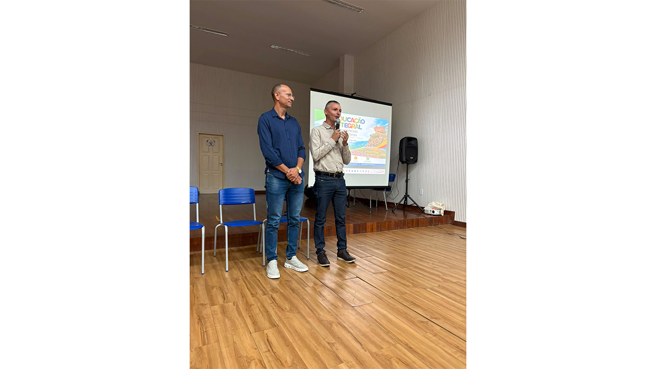 Dois homens fazem uma apresentação em uma sala com piso de madeira. O homem da direita, de camisa bege, fala ao microfone. O homem da esquerda veste camisa azul escura e óculos. Ao fundo, uma tela de projeção com o texto "Educação Integral" e três cadeiras plásticas azuis vazias.
