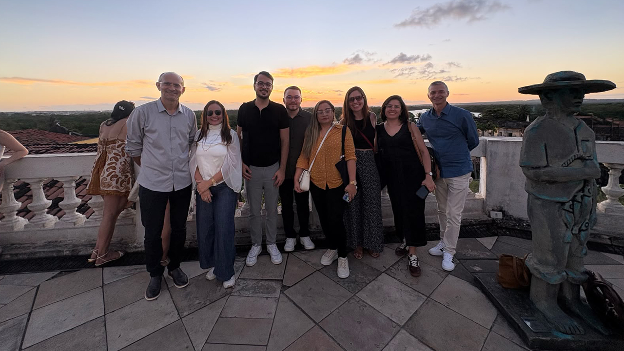 Um grupo de oito adultos posa em pé em um terraço de pedra ao pôr do sol. O céu tem cores laranja e azul. O grupo sorri para a câmera com vestimentas casuais. À direita, uma escultura de pedra escura de uma figura humana com chapéu. Atrás das pessoas, há um guarda-corpo em pedra na cor branca.
