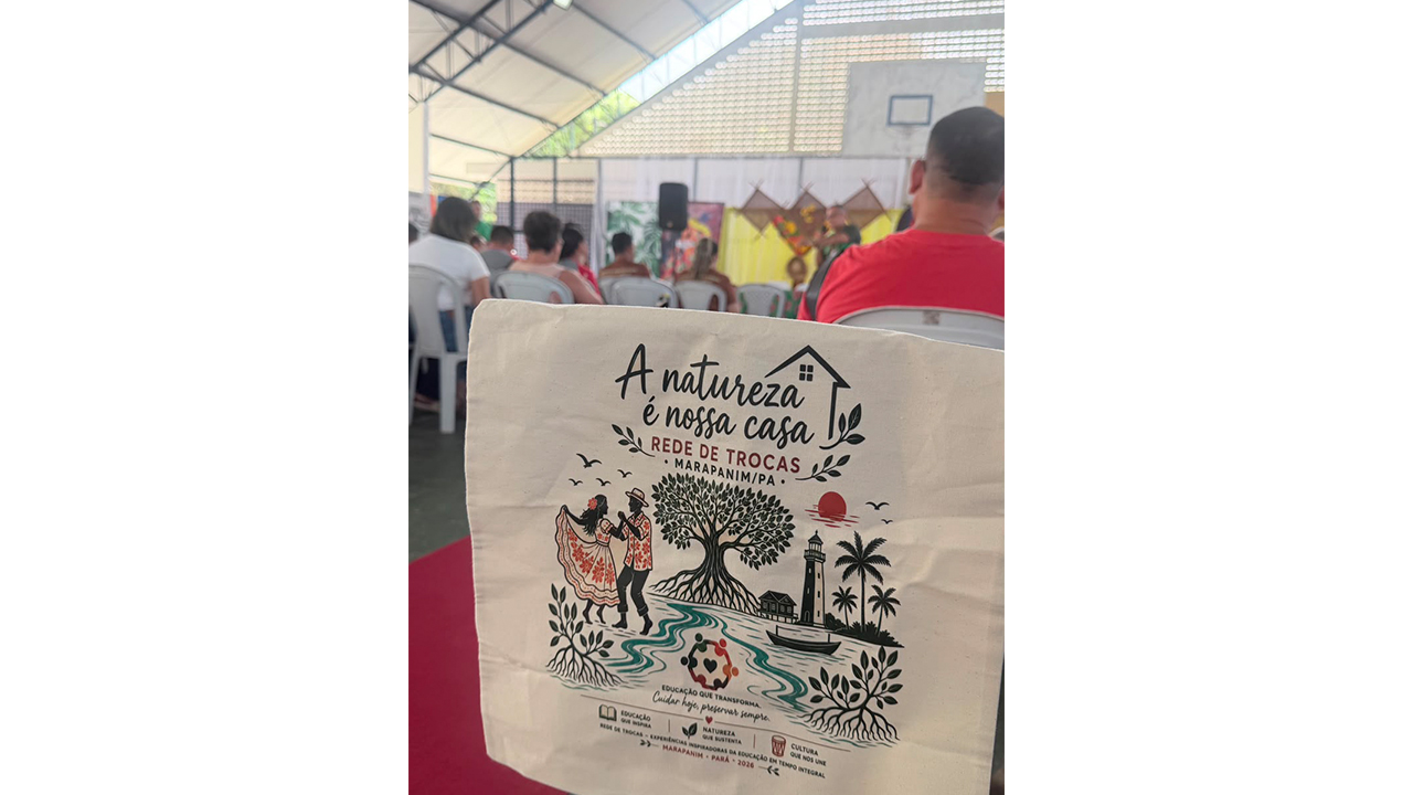 Várias pessoas estão sentadas em cadeiras brancas de plástico, observando uma apresentação ao fundo. O principal elemento em primeiro plano é uma ecobag de tecido claro, que exibe a ilustração de uma árvore, um casal dançando e os textos “A natureza é nossa casa” e “Rede de Trocas Marapanim/PA”.