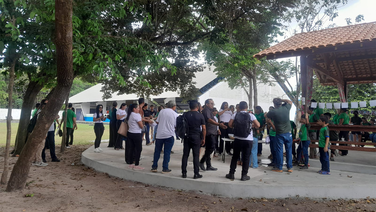 Um grupo de cerca de trinta pessoas, entre adultos e crianças, está em pé, conversando. Todas estão em um espaço ao ar livre, ao lado de uma grande estrutura coberta, feita de madeira e telhas marrons. Há também uma grande árvore cobrindo parte do chão cinza de concreto onde as pessoas estão. À esquerda e ao fundo, há uma construção branca com teto inclinado. O chão é de grama e de terra batida.