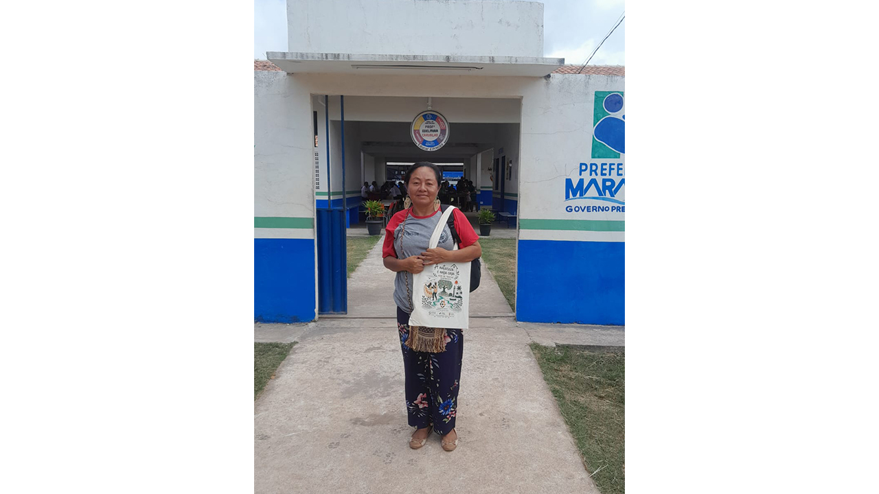 Uma mulher vestindo blusa cinza e vermelha e calça estampada posa em frente à entrada de um prédio térreo. Ela segura uma ecobag clara com ilustrações e a frase “A natureza é nossa casa”. Ao fundo, observa-se a fachada branca com detalhes azuis, um portão e letreiros institucionais.