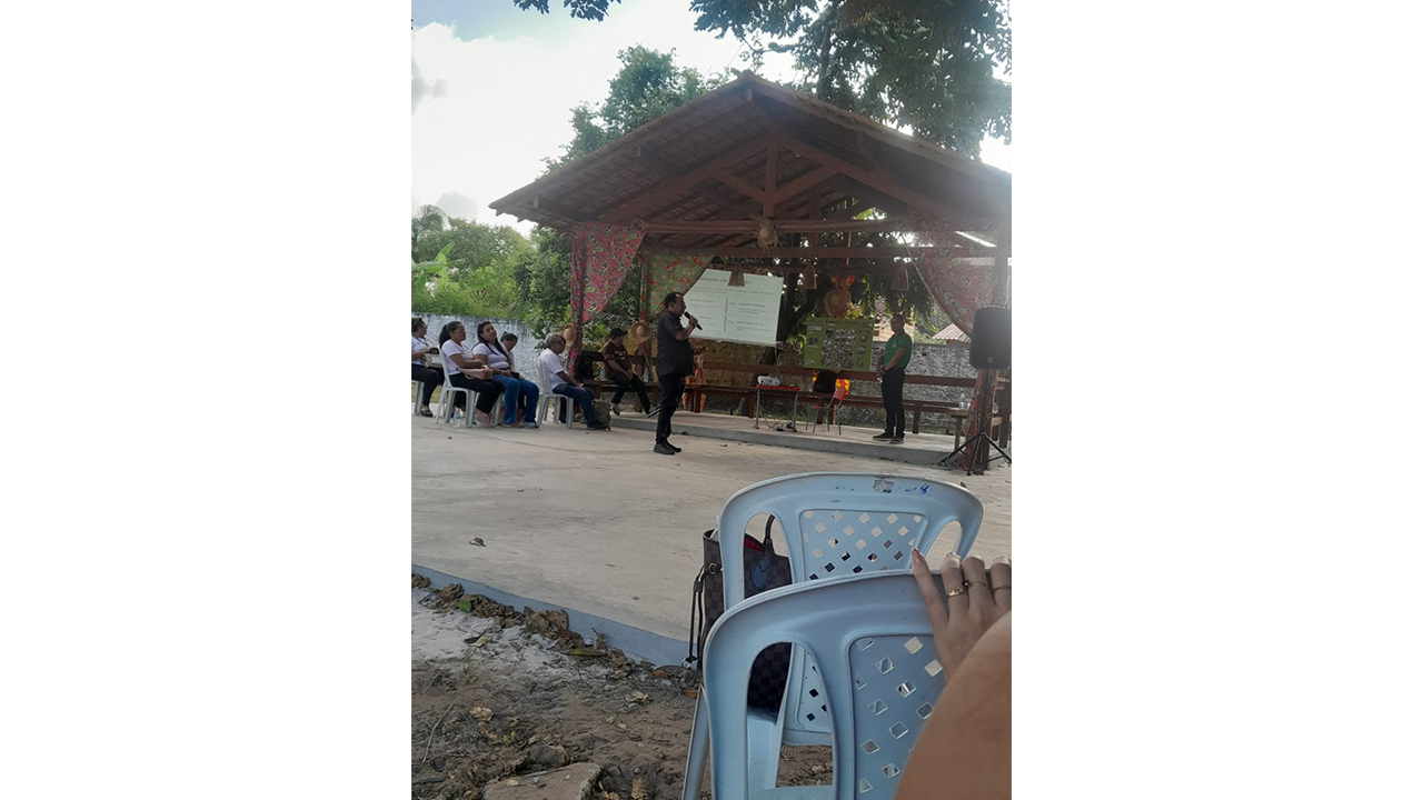 Um homem vestido de preto está em pé, segurando um microfone e gesticulando enquanto discursa para um pequeno grupo sentado em cadeiras plásticas brancas. Atrás dele, um telão projeta gráficos, posicionado em uma tenda rústica de madeira decorada com tecidos florais. Ao fundo, observam-se árvores e o chão de cimento batido.