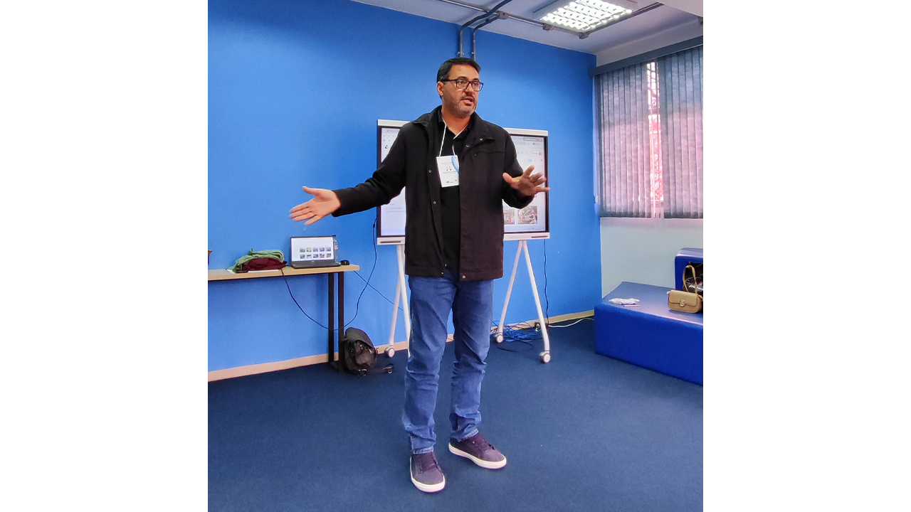 No centro de uma sala de piso e paredes azuis, um homem de jaqueta preta e calça jeans está em pé, falando e gesticulando. Ele usa um crachá e está posicionado em frente a uma tela, montada em um suporte.