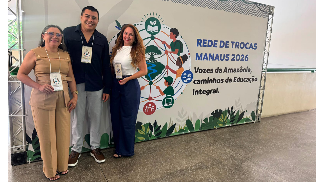 Três adultos posam diante de um painel. Duas mulheres e um homem usam crachás e roupas casuais. O painel exibe “Rede de Trocas Manaus 2026 – Vozes da Amazônia, Caminhos da Educação Integral”, com ilustrações de pessoas e elementos da natureza.