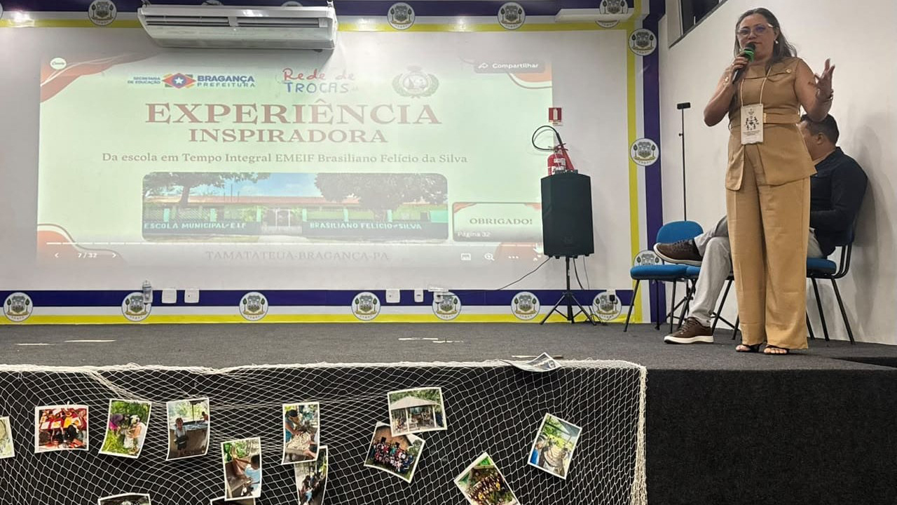 Auditório com uma apresentação projetada ao fundo. Uma mulher vestindo roupa bege fala ao microfone, em pé, enquanto um homem permanece sentado ao lado. Na projeção, lê-se “Experiência Inspiradora”, referente à escola em tempo integral. Fotografias estão presas a uma rede diante do palco.