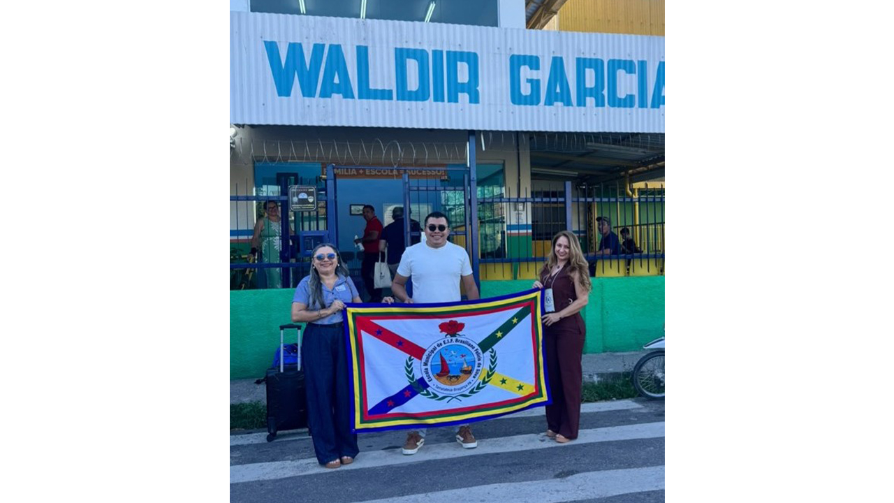 Duas mulheres e um homem posam, em cima da faixa de pedestres na entrada de uma escola, segurando uma bandeira colorida com fundo branco, um brasão ao centro, e linhas formando um 'x', nas cores vermelha, verde, azul e amarela, com uma rosa no topo do brasão circular. O muro da escola é verde, com grades azuis, e acima, em um fundo branco, lê-se parcialmente o nome da instituição em azul: "Waldir Garcia".
