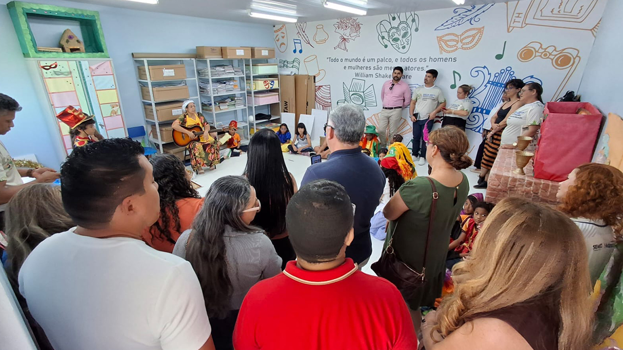 Sala de atividades com adultos e crianças de diferentes perfis raciais reunidos em torno de uma apresentação cultural. Uma mulher sentada toca violão, enquanto crianças com roupas coloridas permanecem próximas e adultos observam. Ao fundo, há estantes e objetos cênicos.