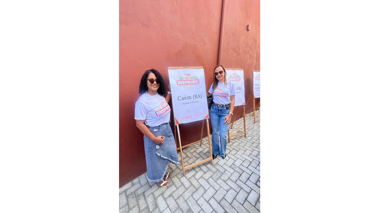 Dupla multirracial de mulheres está em pé, sorrindo e usando óculos escuros, camisetas brancas iguais e calça e saia jeans. Entre elas, um banner em um cavalete de madeira clara exibe o texto “Caém (BA) — Jéssica e Daniela”. Ao fundo, há uma parede lisa vermelho-escura, além do piso de blocos cinza.