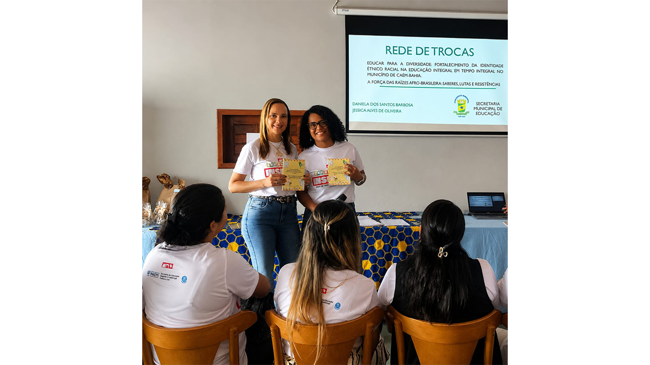 Dupla multirracial de mulheres sorri, em pé, segurando certificados, enquanto é observada por três pessoas de costas, sentadas em cadeiras de madeira. Ao fundo, um telão projeta o texto “Rede de Trocas”, os nomes "Daniela e Jéssica" e o título "Educar para a diversidade: fortalecimento da identidade étnico-racial na Educação Integral em Tempo Integral no município de Caém (BA)". O cenário exibe paredes brancas, cadeiras de madeira e uma longa mesa com uma toalha estampada nas cores azul marinho e amarelo.