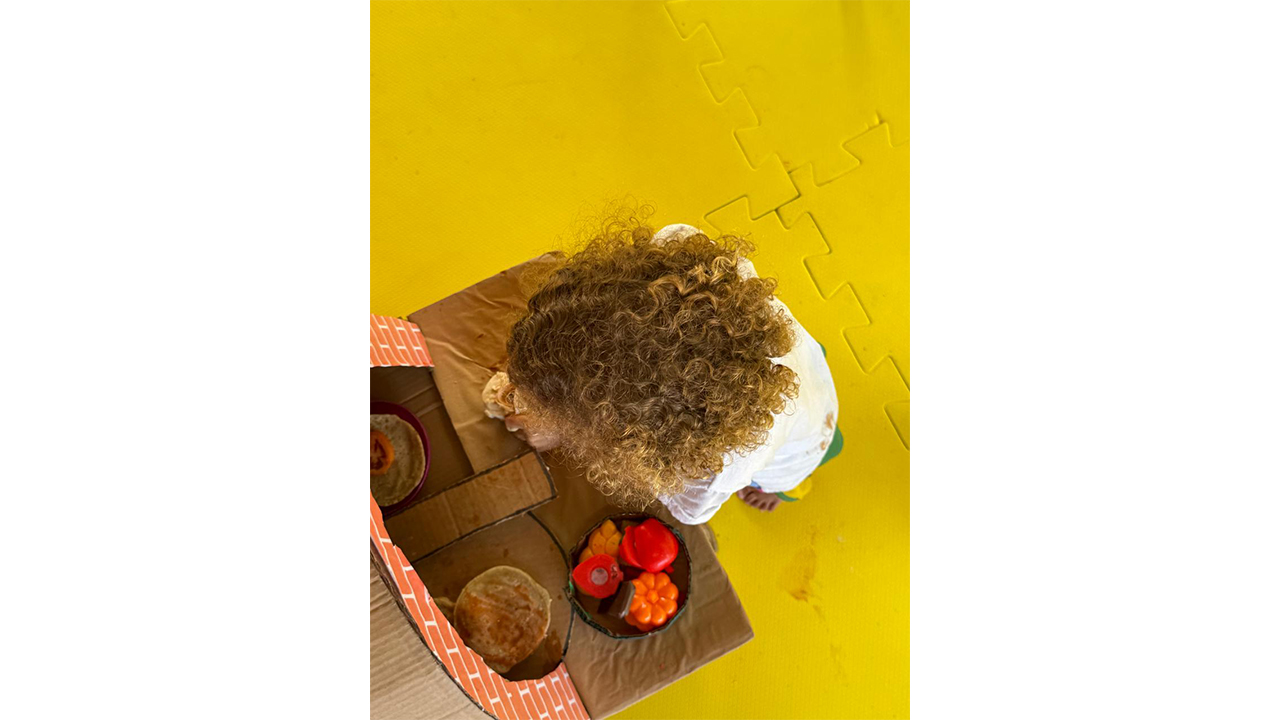 Uma criança, vista de cima, brinca sobre um tapete de borracha amarelo. O destaque é uma estrutura de papelão, com um forro de tijolos imitando um forno, com pratinhos com alimentos de plástico.