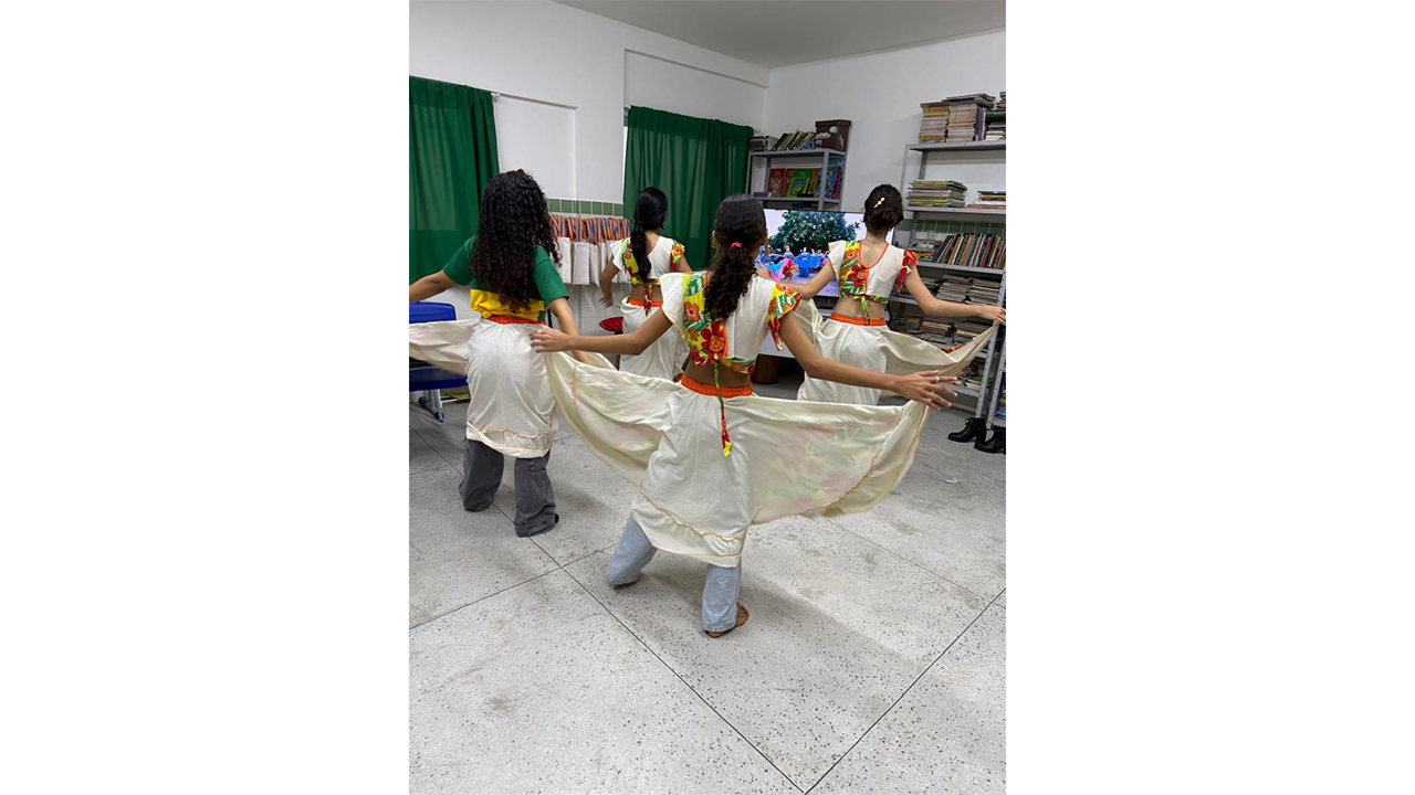 Grupo multirracial de quatro jovens, visto de costas, dança segurando as bordas de longas saias brancas, combinadas com blusas curtas com estampas florais. À frente, uma televisão exibe um vídeo de dança semelhante. O grupo está em uma sala com piso claro, cortinas verdes e uma estante de metal cheia de livros à direita.