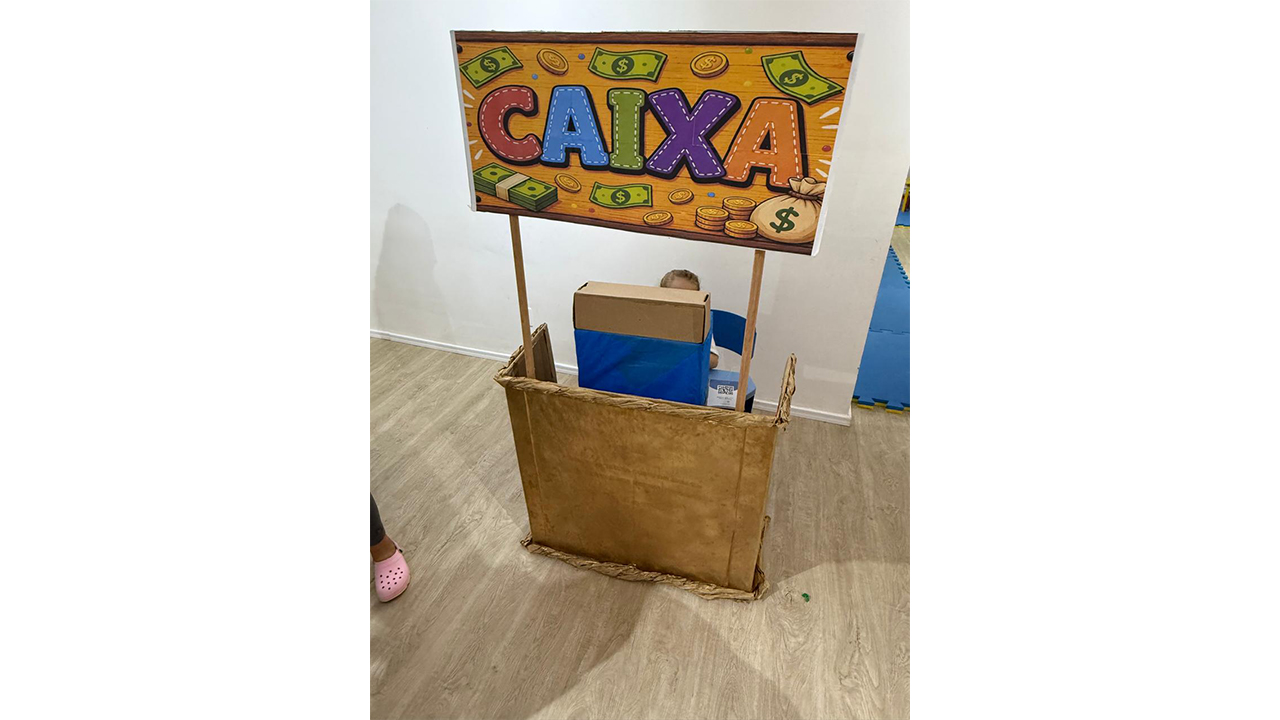 Uma estrutura de papelão imita um balcão, com caixas empilhadas e uma placa superior com a palavra "CAIXA", escrita em letras coloridas e decorada com desenhos de notas e moedas. A sala conta com piso laminado de madeira clara e parede branca.