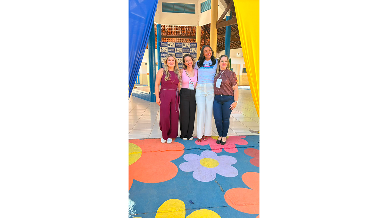 Um grupo multirracial de quatro mulheres posa para foto em um pátio escolar. Elas estão lado a lado, com os braços próximos umas das outras e sorrindo, com roupas casuais e algumas com crachás de identificação. O espaço possui colunas azuis, tecidos amarelos e azuis e piso pintado de azul com grandes flores coloridas: laranja, rosa, lilás e amarela. Ao fundo, em uma parte coberta, o piso é de cerâmica branca.