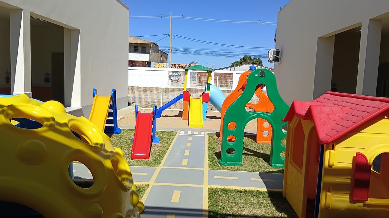 Área recreativa externa de uma unidade escolar. O espaço possui escorregadores, estruturas de escalada e uma casinha infantil nas cores verde, amarelo, vermelho, azul e laranja. No chão, há gramado e um percurso pintado em cinza com faixas amarelas e de pedestre que simula uma pequena via, além de um escorregador em um trecho de areia. Nas laterais da imagem, é possível ver a estrutura branca da escola, e, ao fundo, um portão branco e a rua, com casas e postes de iluminação.