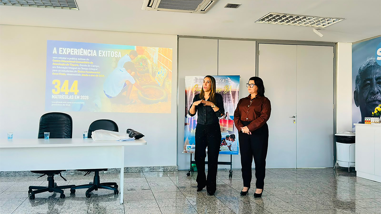 Em uma sala, duas mulheres estão em pé diante de uma projeção, fazendo uma apresentação. Na tela, lê-se "A experiência exitosa" e "344 matrículas em 2026", juntamente com a foto de duas crianças agachadas no canto direito da tela. À frente, há uma mesa com copos d'água, duas cadeiras pretas, um banner e uma porta dupla branca fechada, juntamente com um painel institucional à direita.