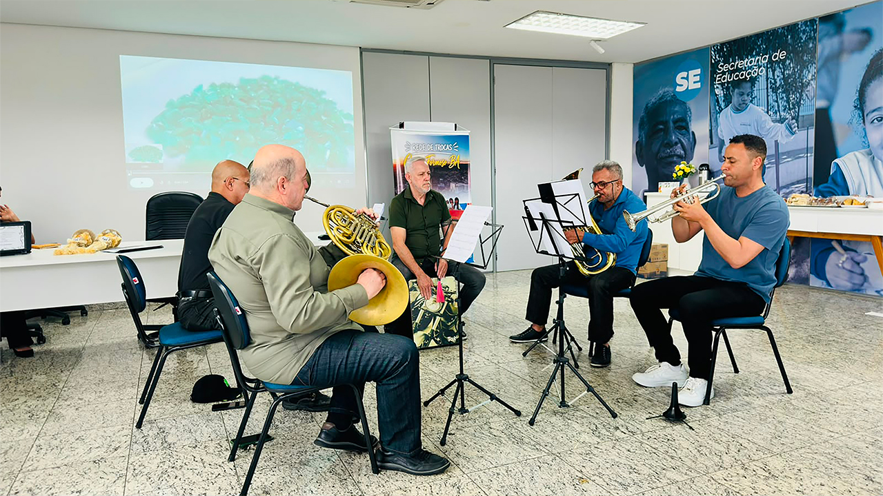 Cinco homens realizam uma apresentação musical, sentados em semicírculo em cadeiras pretas com estofado azul. Eles tocam instrumentos de sopro, como trompete e trompas, diante de estantes pretos com partituras. Ao fundo, em um painel institucional azul, aparecem os textos "Secretaria de Educação", "SE" e um banner de Campo Formoso com o título "Rede de Trocas".