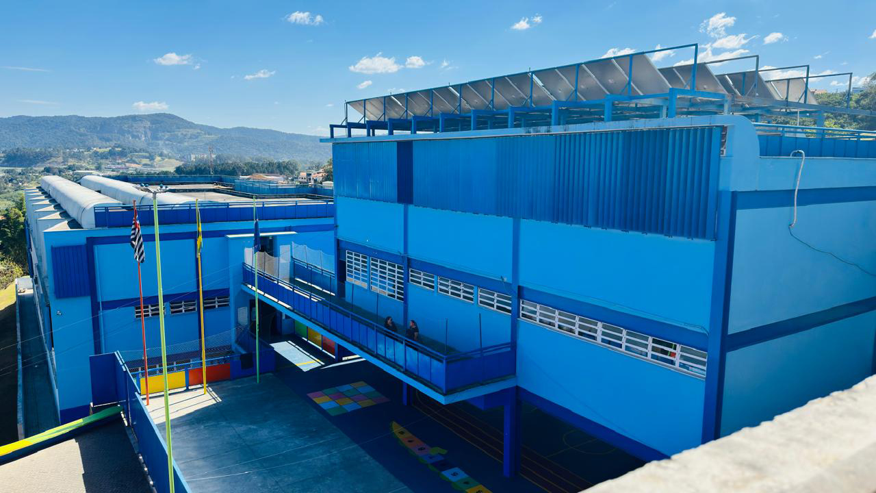 Vista externa de uma edificação escolar de grande porte, predominantemente azul. No pátio, há mastros com bandeiras, rampas e marcações coloridas no piso. Já no último piso, há quatro fileiras de painéis solares. Ao fundo, aparecem outras construções, vegetação e montanhas sob o céu azul. A foto foi tirada do alto de outra construção.