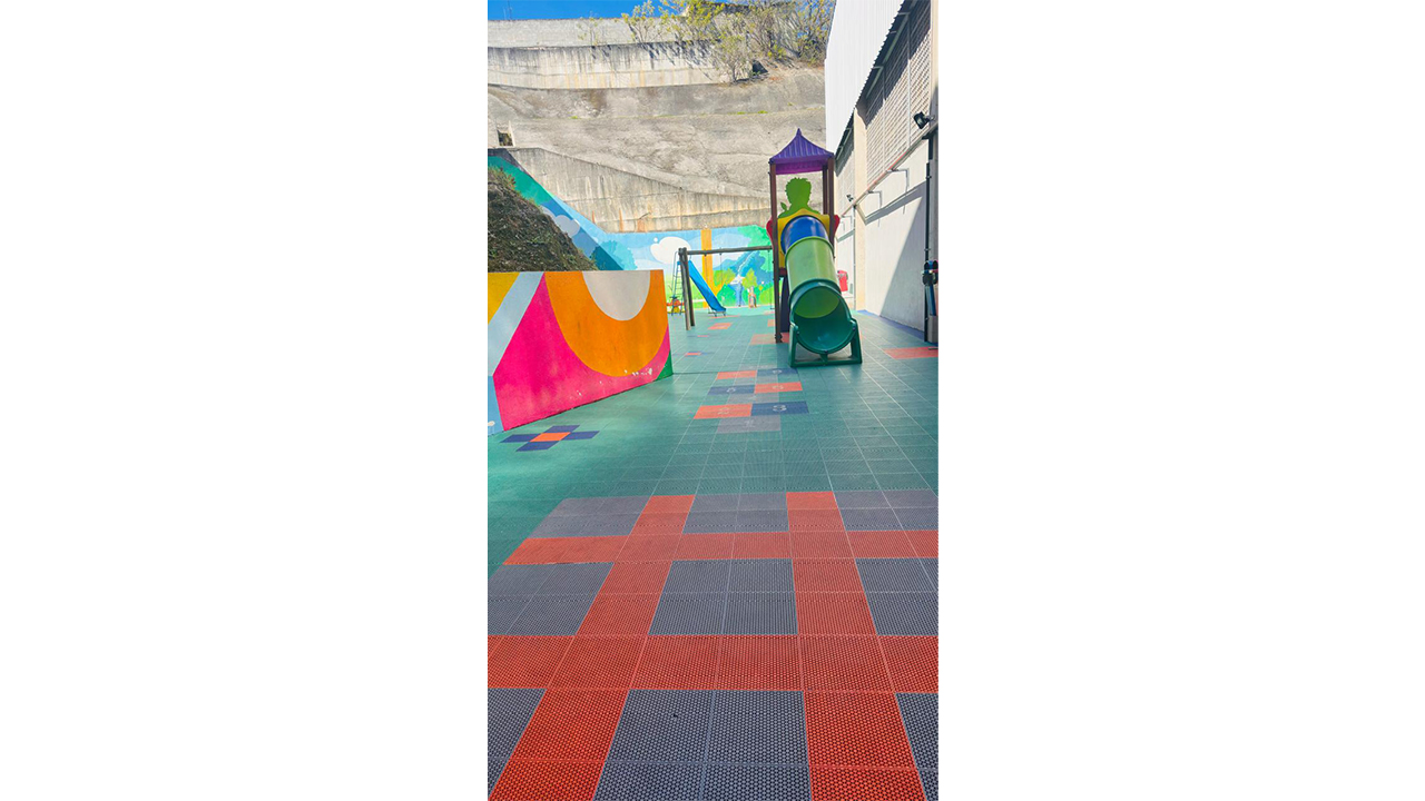 Área recreativa externa com brinquedos infantis coloridos e de plástico e piso modular colorido. Ao centro, há um escorregador tubular verde e, mais ao fundo, um balanço e outro escorregador azul. As paredes apresentam pinturas geométricas coloridas. Ao fundo, um muro cinza delimita o espaço.