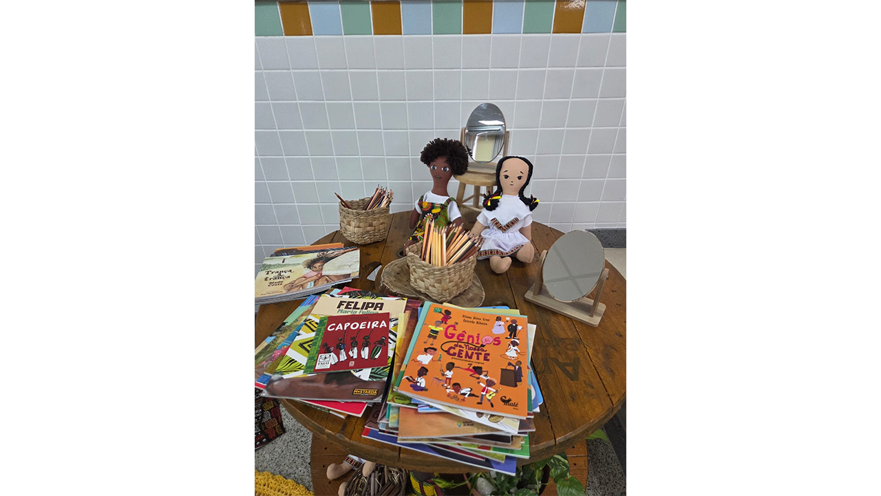 Uma mesa de madeira redonda exibe livros infantis empilhados, acompanhados de duas bonecas de pano, pequenos espelhos de mesa e cestos de palha com lápis de cor. O plano de fundo é composto por uma parede revestida com azulejos brancos, intercalados por uma faixa de azulejos verdes e laranjas.