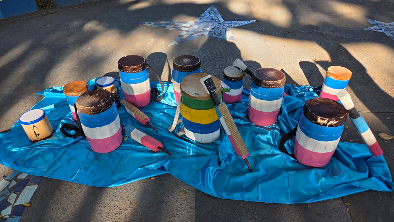 Um conjunto de instrumentos, como tambores e baquetas listradas nas cores azul, branca, rosa, verde e amarela, está disposto no chão, sobre um tecido azul. O plano de fundo mostra um piso de cimento cinza, decorado com estrelas de mosaico branco e azul.