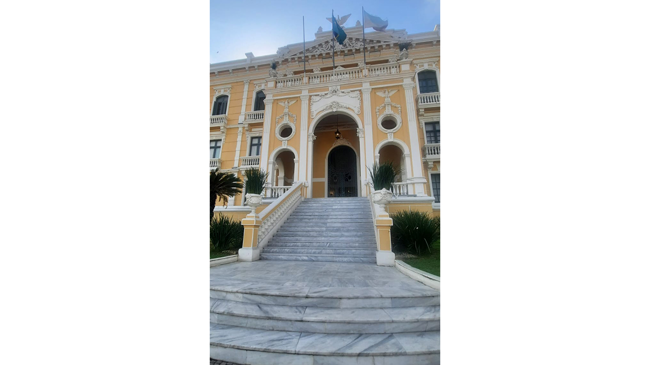 Fachada de um grande edifício histórico sob um céu azul. Uma escadaria reta conduz a uma entrada principal em arco. O plano de fundo revela a arquitetura do prédio, pintado de amarelo, com ornamentos e colunas em branco, vasos de plantas verdes e calçamento de pedras na base.