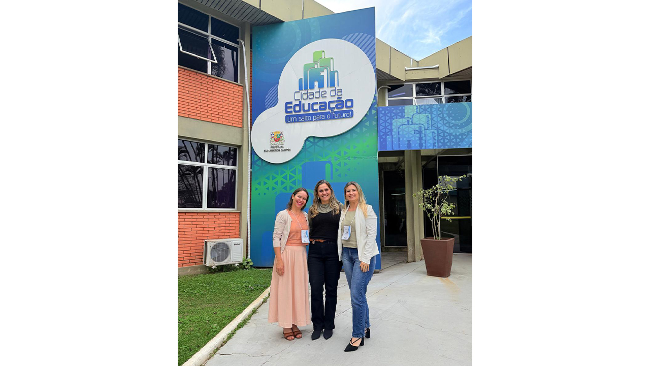 Três mulheres, em trajes casuais, estão em pé, lado a lado, sorrindo em frente à fachada de um edifício com o letreiro “Cidade da Educação - Um salto para o futuro!” e o logotipo da Prefeitura de São José dos Campos. O prédio é de tijolos, com janelas escuras de vidro, enquanto o painel com o letreiro é azul com tons de verde e o letreio está em um círculo branco.