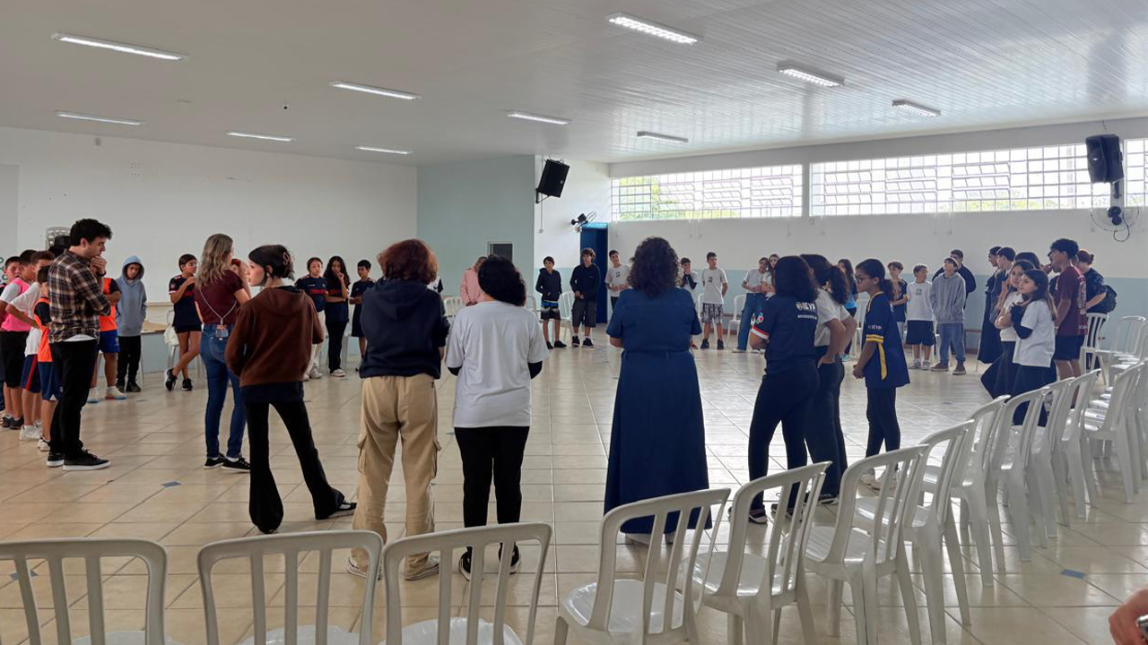 Um grupo multirracial de cerca de cinquenta pessoas, entre adultos e crianças, está em um salão. Todos estão em pé, formando uma roda voltada para o centro. Em primeiro plano, cadeiras de plástico brancas vazias estão organizadas em círculo. O espaço tem paredes brancas, piso de cerâmica clara e janelas ao fundo.