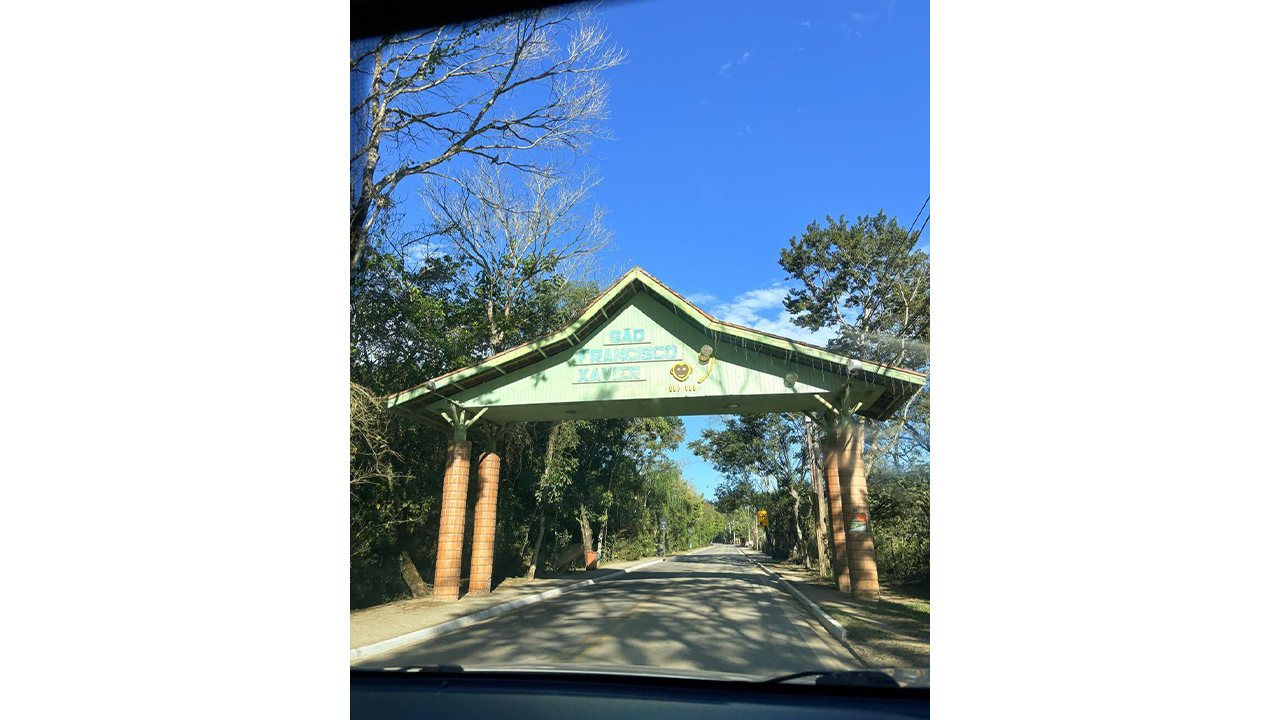 Um portal de madeira verde está sobre a via, sustentado por pilares de tijolos, exibindo o texto "SÃO FRANCISCO XAVIER". É possível ver a rodovia asfaltada, contornada por densa vegetação verde, sob um céu azul.
