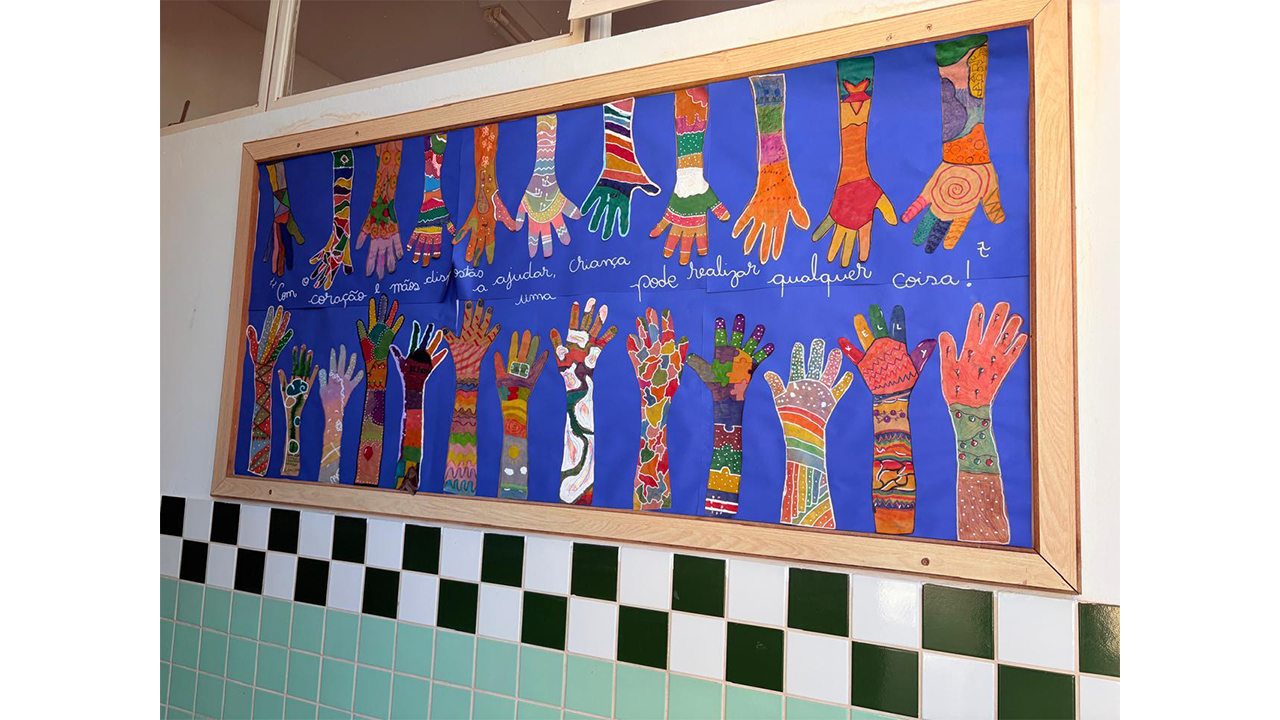 Um mural decorativo está fixado em uma parede. Com um fundo azul, exibe dezenas de recortes de mãos e braços pintados com desenhos e padronagens coloridas, além da frase manuscrita "Com o coração e mãos dispostas a ajudar, uma criança pode realizar qualquer coisa!". Abaixo do mural, há uma parede com azulejos em tons de verde, preto e branco.