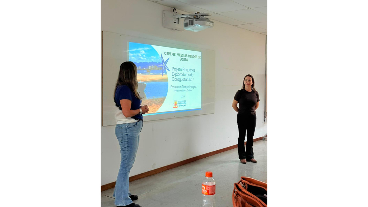 Em uma sala interna de paredes brancas, duas mulheres estão em pé em frente a uma tela de projeção, que exibe a foto de uma praia e o texto "CEI/EMEI Messias Mendes de Souza - Projeto pequenos exploradores de Caraguatatuba". Elas vestem roupas casuais, como jeans e camisetas.