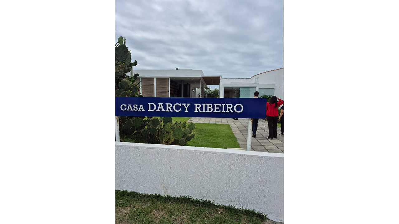 Em primeiro plano, uma placa azul exibe o nome da instituição. Ao fundo, há uma edificação branca, jardim com gramado e cactos e algumas pessoas próximas à entrada.