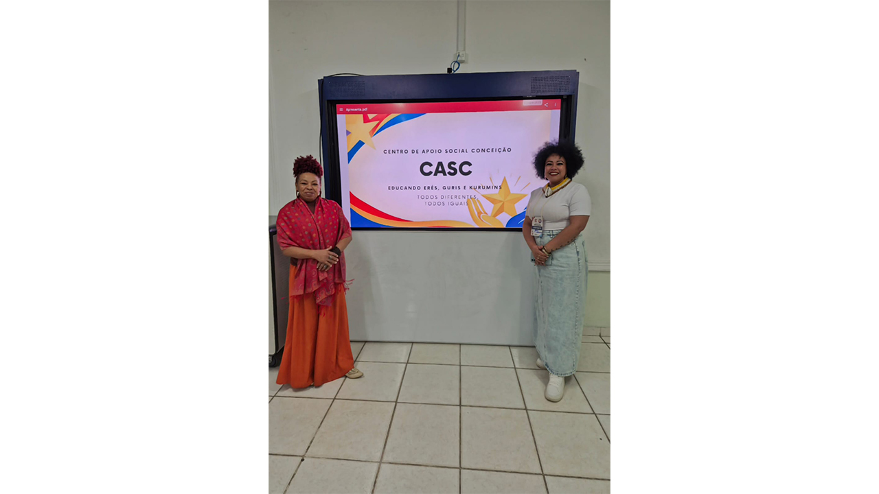 Ambiente interno com duas mulheres em pé diante de uma tela digital. Na tela, lê-se “Centro de Apoio Social Conceição – CASC” e “Todos diferentes, todos iguais”. Uma mulher veste tons de vermelho e laranja, e a outra usa camiseta clara e saia jeans.