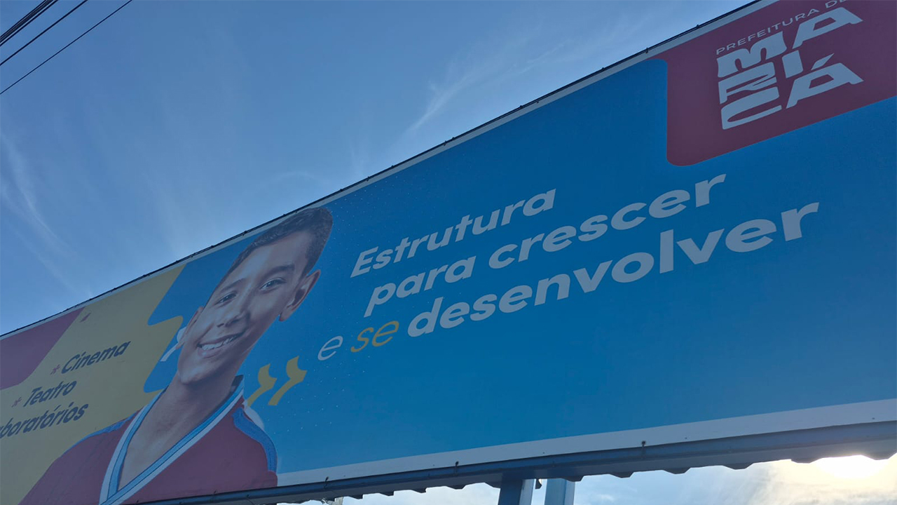 Área externa com grande painel institucional. O painel apresenta a imagem de uma criança e a frase “Estrutura para crescer e se desenvolver”, além da identificação da Prefeitura de Maricá. Na lateral, aparecem referências a cinema, teatro e laboratórios.