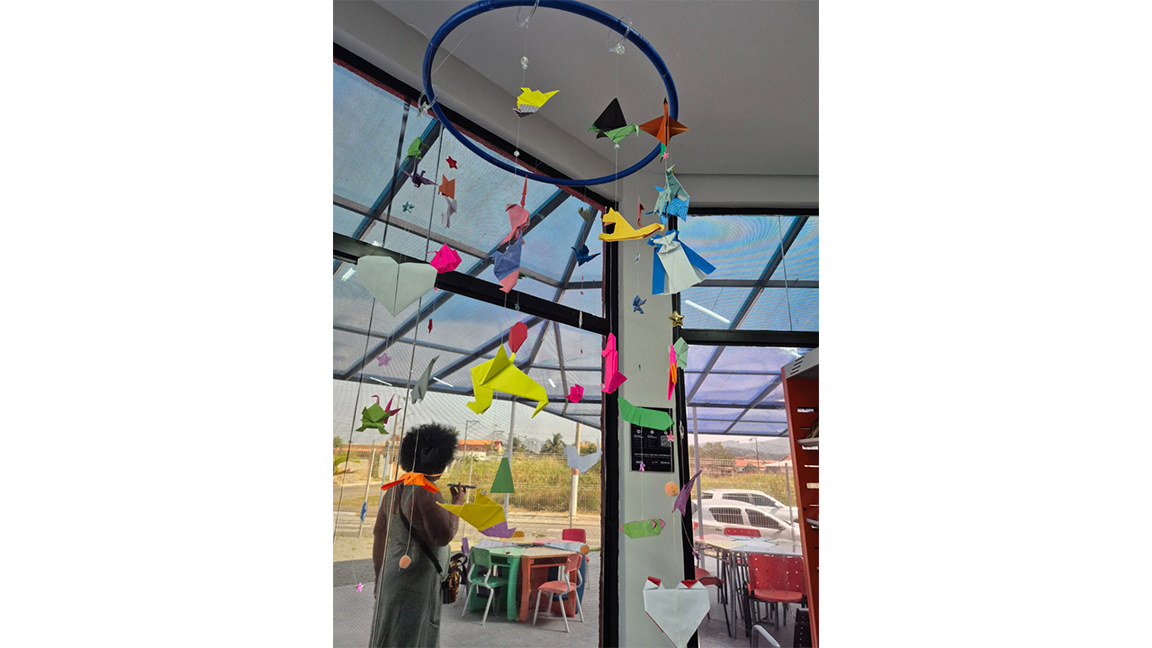 Espaço interno decorado com móbiles de origami suspensos em um aro azul. As dobraduras têm diferentes formas e cores e ficam diante de grandes janelas. Ao fundo, há mesas e cadeiras e uma pessoa em pé próxima à área externa.