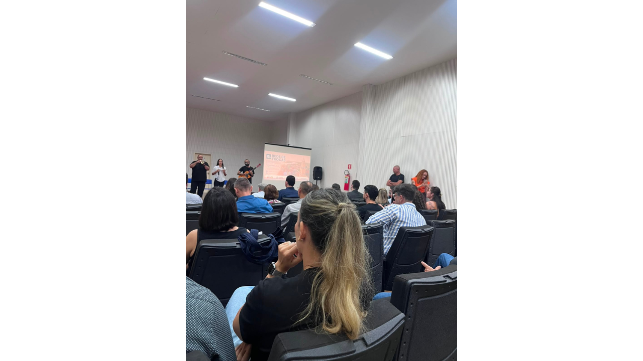 Em uma apresentação, pessoas sentadas em cadeiras pretas de plástico observam um trio no palco: à esquerda, um homem toca percussão; ao centro, uma mulher segura um microfone; e, à direita, um homem toca violão. Atrás do trio, um telão projeta uma imagem com o título "Rede de Trocas". O ambiente possui paredes e teto branco.
