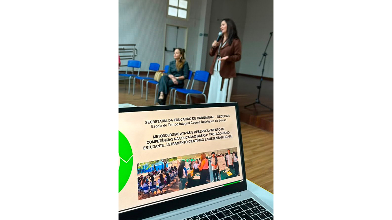 Em um auditório, um notebook sobre uma mesa exibe uma imagem de apresentação. Ao fundo, duas mulheres: uma, de blazer marrom, fala ao microfone de pé, enquanto a outra está sentada, usando roupas casuais e com as pernas cruzadas. O espaço conta com um palco de madeira, cadeiras azuis vazias e uma parede branca, além de máquinas de ar condicionado próximas ao teto branco.