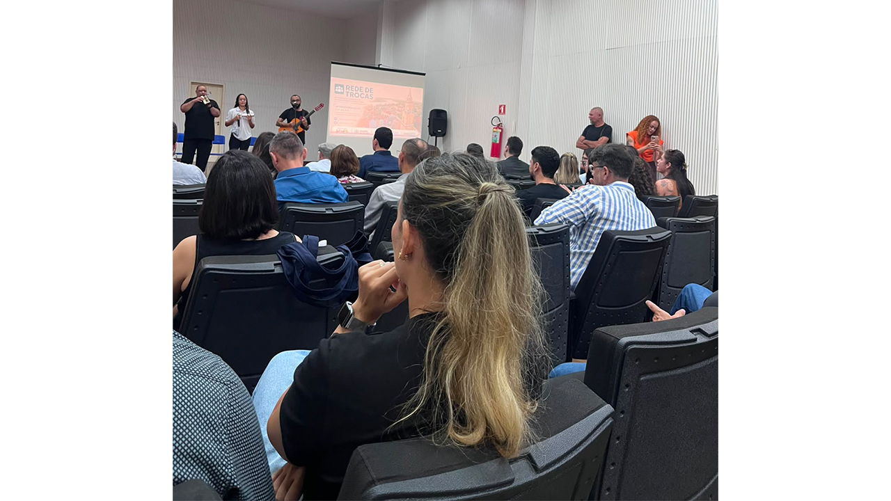 Em uma apresentação, pessoas sentadas em cadeiras pretas de plástico observam um trio no palco: à esquerda, um homem toca percussão; ao centro, uma mulher segura um microfone; e, à direita, um homem toca violão. Atrás do trio, um telão projeta uma imagem com o título "Rede de Trocas". O ambiente possui paredes e teto branco.