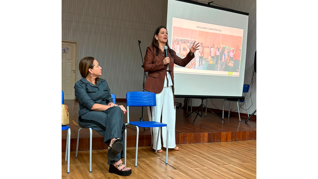 No centro de um auditório, uma mulher em pé, de blazer marrom e calça branca, gesticula enquanto fala ao microfone, enquanto outra mulher, de conjunto cinza, a observa sentada, com as pernas cruzadas. Em um telão à sua direita, uma apresentação é exibida. O piso é de madeira, há cadeiras plásticas azuis e uma parede branca com frisos verticais.