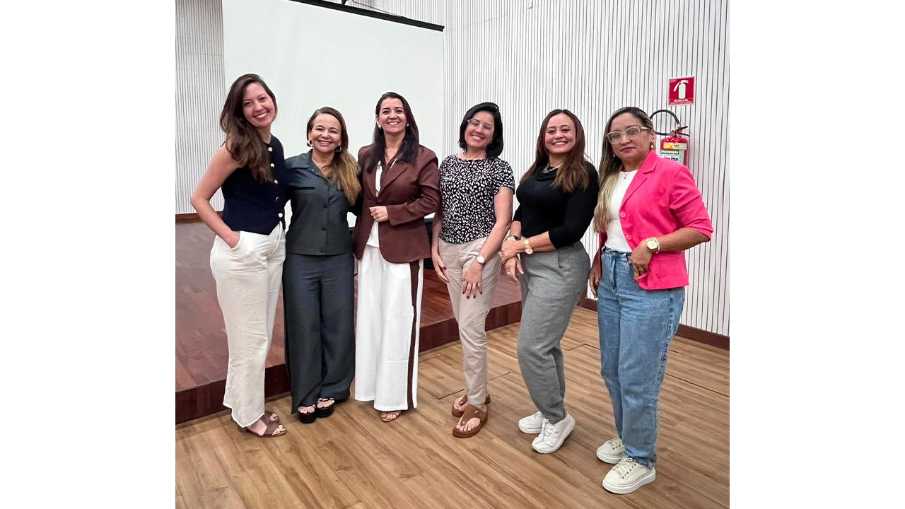 Seis mulheres posam em pé, lado a lado, e sorriem para a câmera, vestindo calças e blusas sociais. Atrás delas, um telão de projeção branco está desligado. O piso é de madeira, as paredes são brancas e ripadas e um extintor de incêndio vermelho está preso à direita.