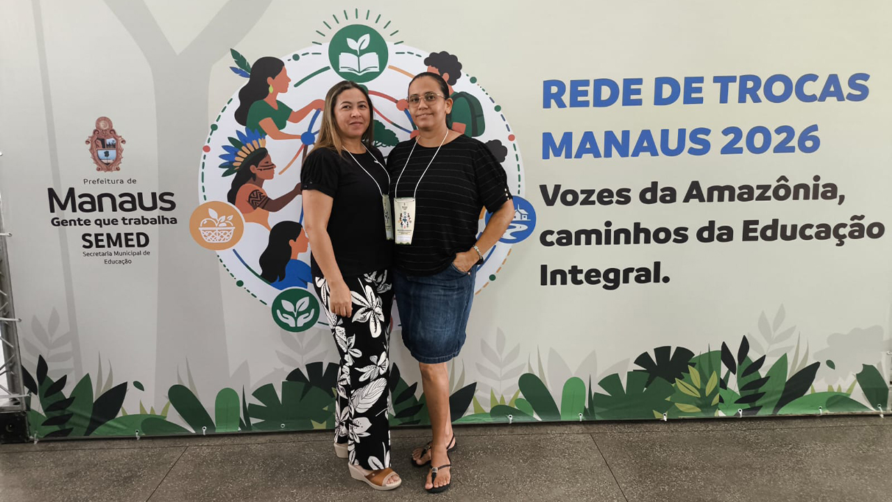 Duas mulheres em pé vestem roupas casuais e usam crachás no pescoço. Elas estão sobre um chão escuro, à frente de um grande painel com o texto "Rede de Trocas Manaus 2026" e ilustrações.