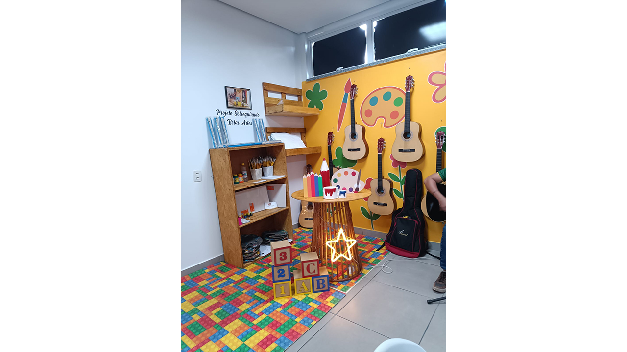Ao centro de uma sala, uma parede amarela exibe três violões pendurados e uma estante com materiais de arte. O chão é coberto por um tapete de EVA colorido, contendo uma mesa central, blocos com letras e uma estrela luminosa.