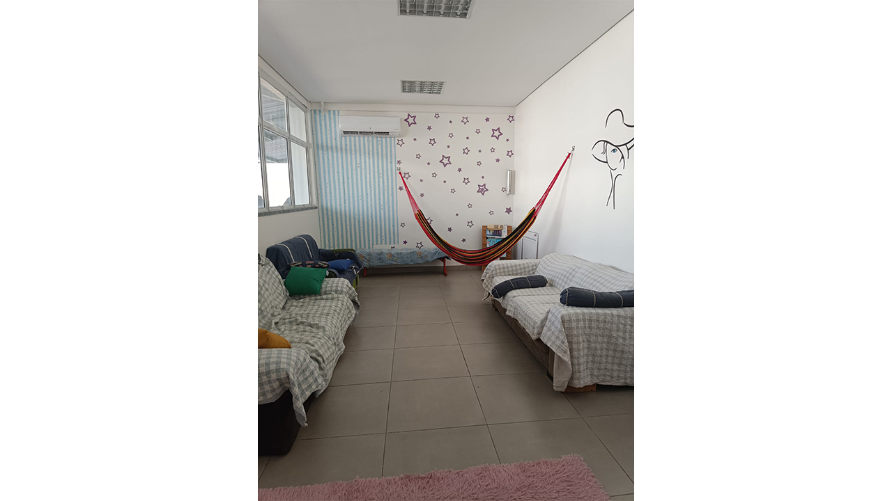 Em uma sala de descanso, dois sofás cobertos por mantas xadrez estão dispostos frente a frente, e uma rede listrada está armada ao fundo. As paredes são brancas, sendo a do fundo decorada com adesivos de estrelas, com piso cerâmico liso e a borda de um tapete rosa visível abaixo.