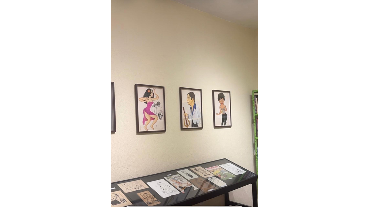 Em uma sala, três quadros na parede exibem caricaturas coloridas, e, abaixo, uma vitrine de vidro exibe jornais antigos. O fundo é composto por uma parede clara.