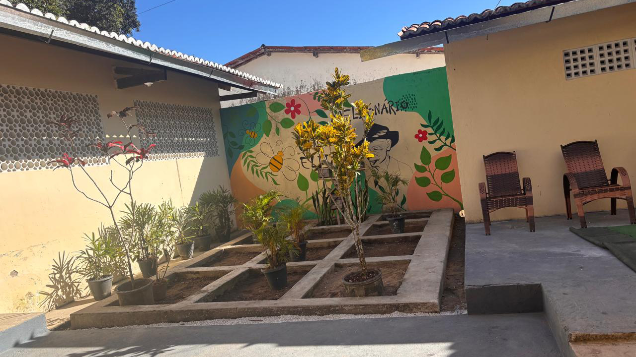 Em um quintal, há um canteiro de cimento dividido em pequenos quadrados, com vasos de plantas sobre a terra batida. À direita, há duas cadeiras de vime e um mural colorido na parede, com um desenho de abelha, uma pessoa de chapéu preto e folhagens. À esquerda, há uma porção de vasos de plantas. Ao redor no canteiro, há construções com paredes amarelas. O chão é de cimento.
